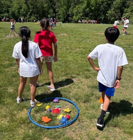 Kids in a grassy field with a hula hoop filled with colorful objects, one wearing a red shirt.