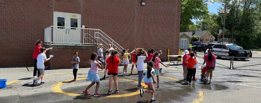 A group of children in red and white shirts dance in a circle on a wet street in front of a brick building.