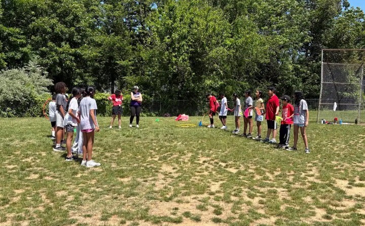 Group of people in casual sports attire stand in a line on a grassy field with trees and a fence.