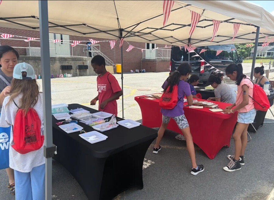 Group of people gather under tent, examining items on tables covered with red and black cloth.