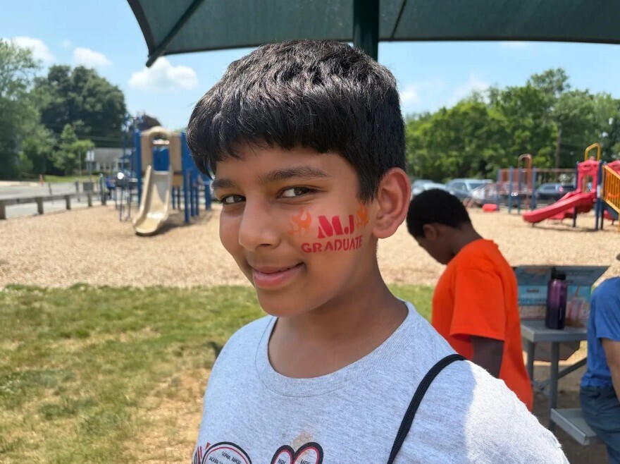 A young boy with face paint stands in a park near playground equipment, wearing a white shirt.