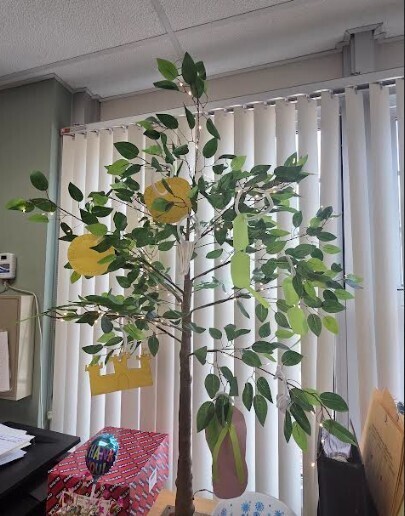 A decorated artificial lemon tree in front of a window with white blinds. Papers and a box are nearby.