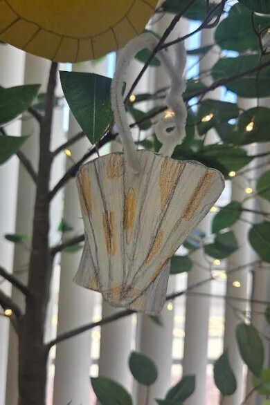 A decorative ornament with a scalloped shell shape hangs from a tree branch. The ornament is white with yellow accents. Green leaves and lights are in the background.