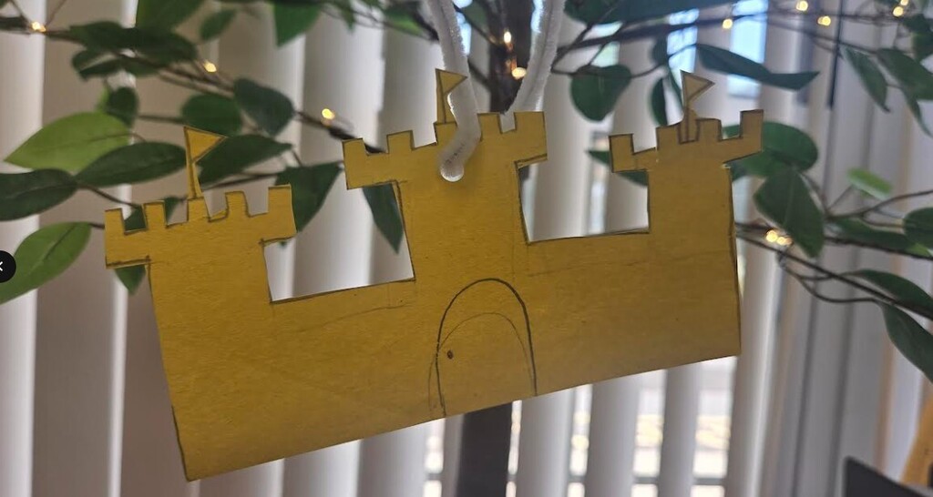 A golden castle paper craft hangs from a tree branch, with green leaves and fairy lights behind a window.