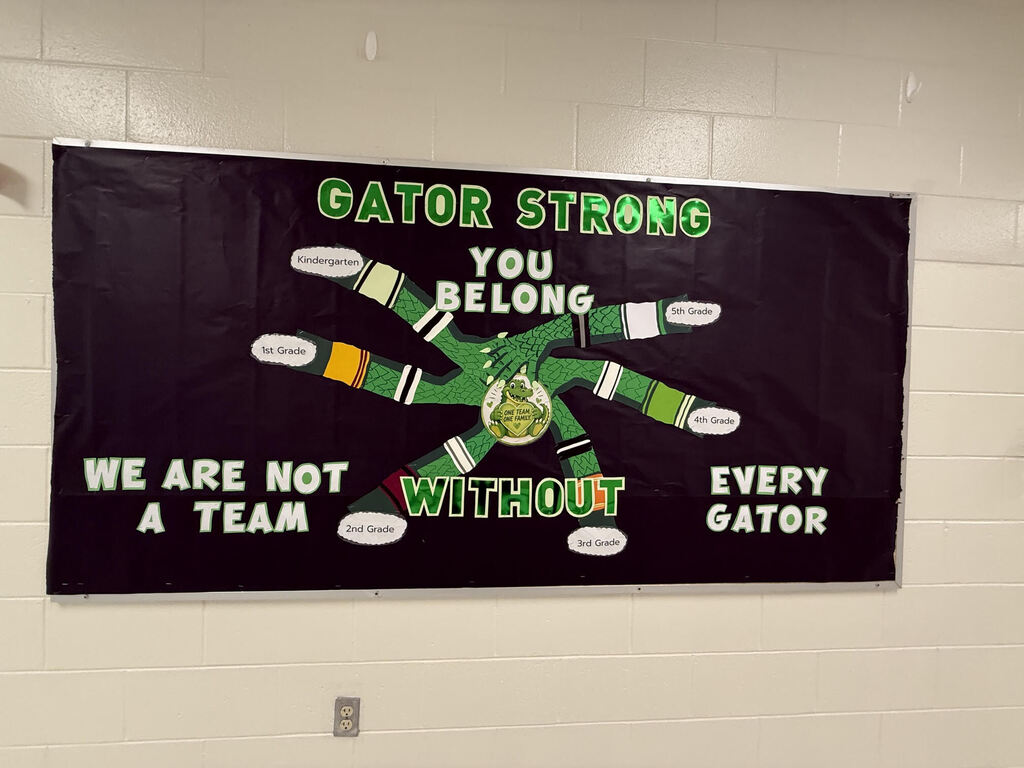 Banner reads "Gator Strong You Belong We Are Not A Team Without Every Gator". White brick background.
