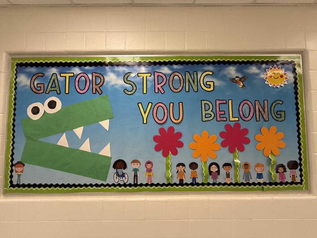 A bulletin board displays a green alligator with eyes, a motivational message, and colorful flowers. A sun is in the sky.