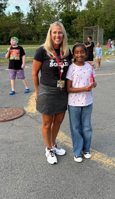 Woman in black shirt with name tag stands with young girl in light pink shirt, both smiling.