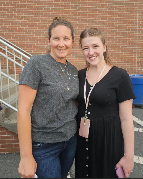 Two women stand together, smiling, outside near stairs. One wears a gray shirt, necklace, and jeans. The other has a black dress and pink lanyard.