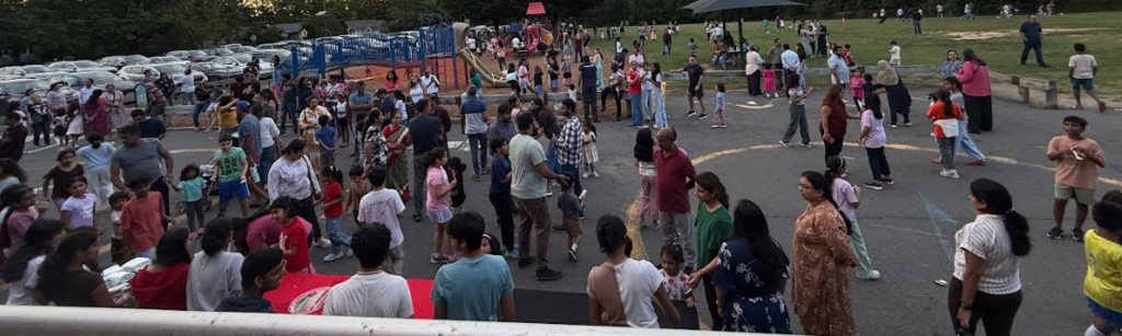 Many people gathered outdoors, some walking, some standing. A playground visible in the background.