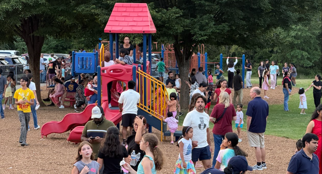 A park with a red slide and climbing structure. Many people, including adults and children, are present.