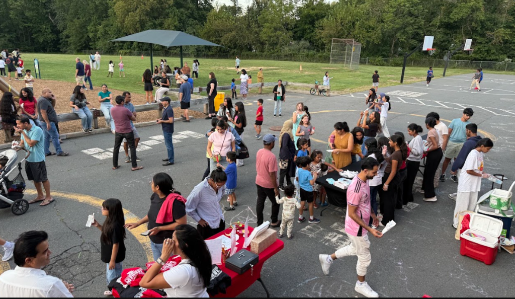 Crowd of people in a parking lot with tables set up, some walking, others standing. Trees and playground in background.