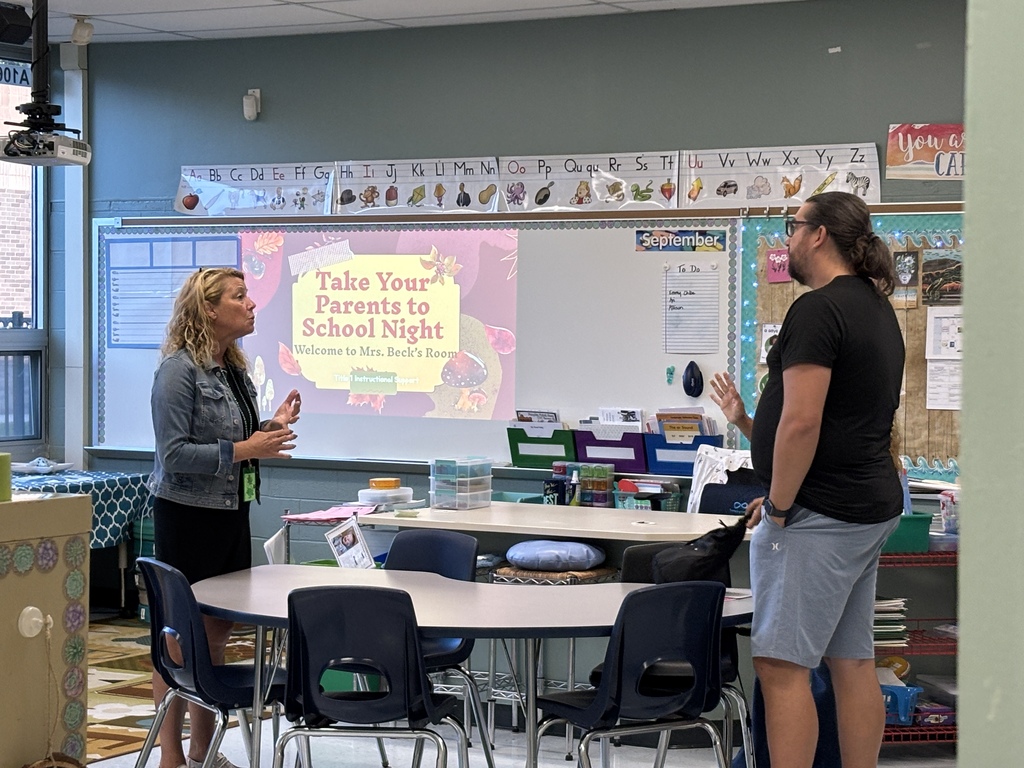 Two individuals are in a classroom with tables, chairs, and a whiteboard displaying a slide titled "Take Your Parents to School Night."
