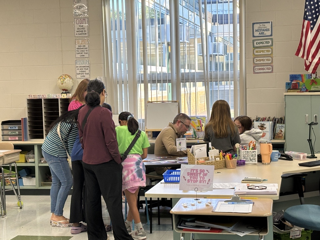 A classroom with desks and shelves. Several people, including adults and children, gather around a table.
