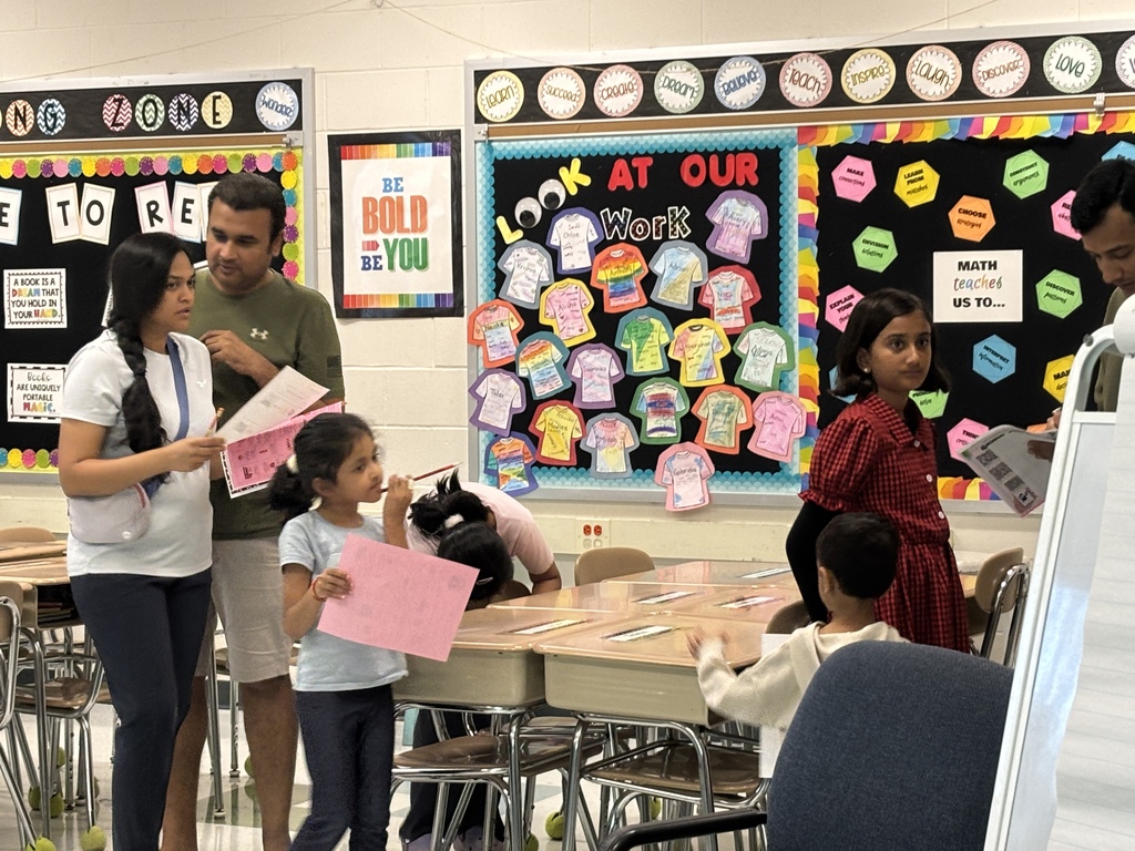 Classroom with students and adults. One woman holds pink paper. A bulletin board has colorful t-shirts.