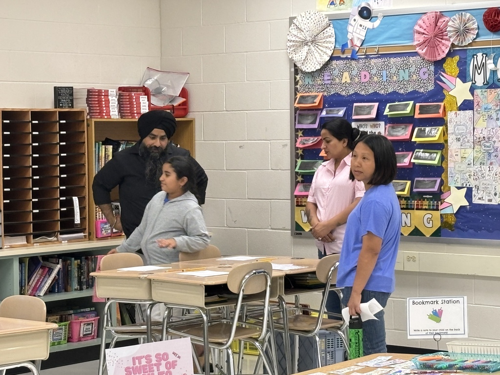 People stand in a classroom with white walls and colorful bulletin boards. Two girls hold papers.