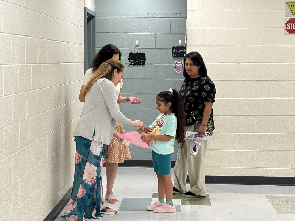A young girl receives a certificate from a woman in a hallway with white walls and a blue floor.