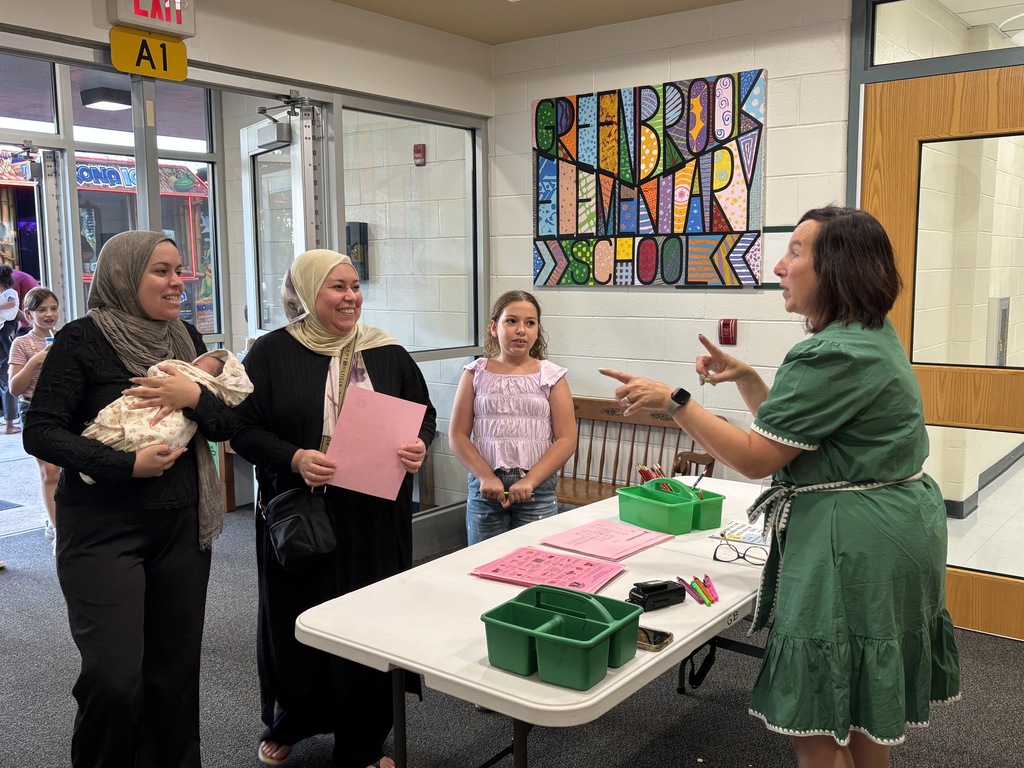 Four people in a hallway, with one woman gesturing, two holding pink papers, and a table with green bins and papers.