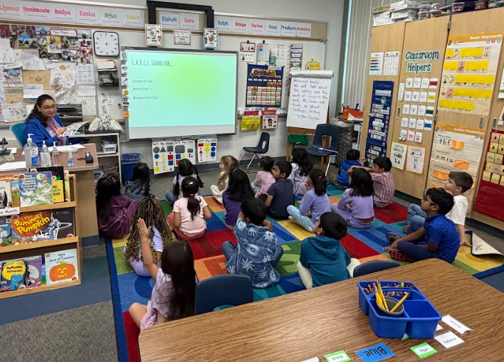 Classroom with students sitting on a rug, a teacher standing by a screen, a whiteboard, and various educational materials.