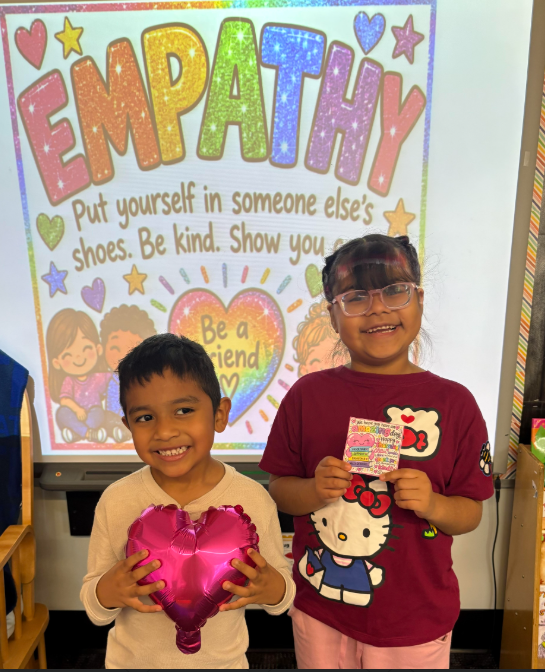 Two children smile and hold cards in front of a classroom screen displaying the word "EMPATHY".