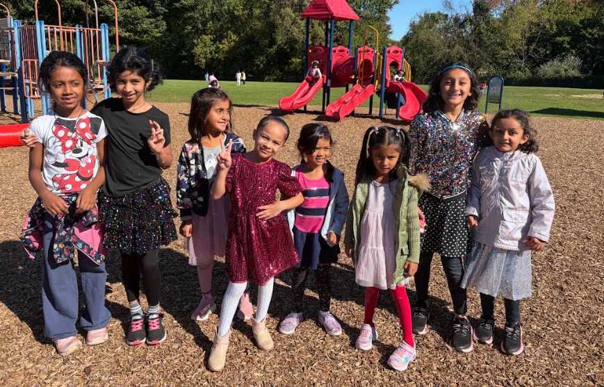 A group of young girls poses in a park. A red and yellow play structure is in the background.