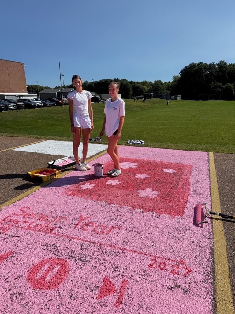 Two women in white uniforms painting a pink square with words and numbers on a paved lot.