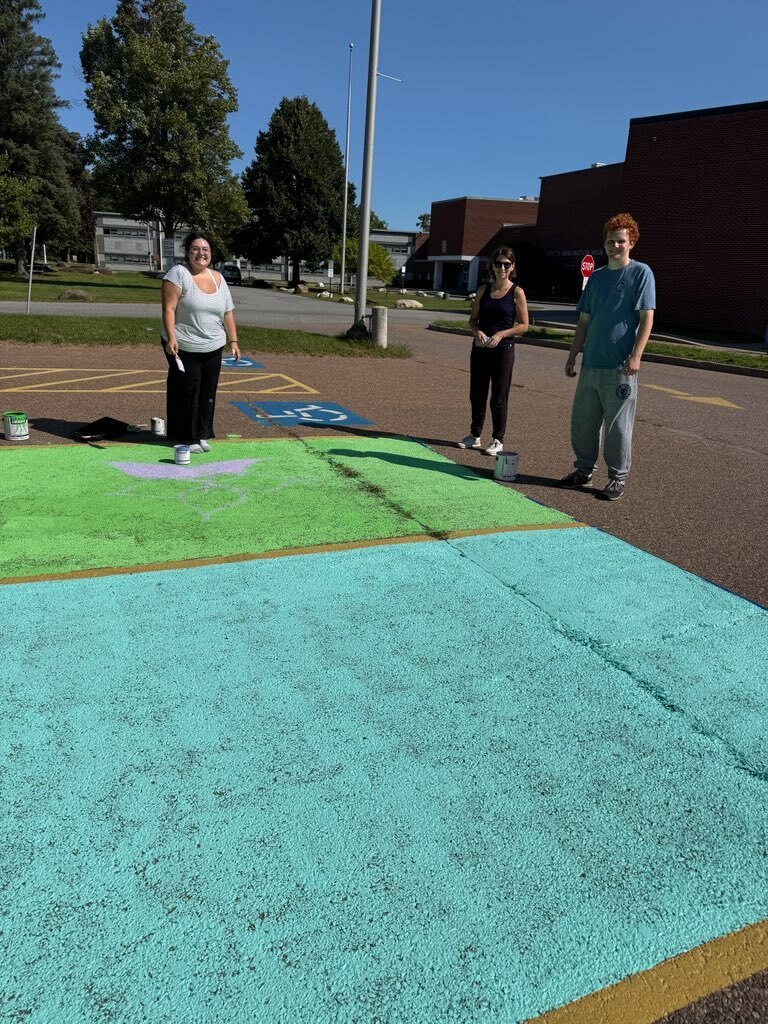 Three people paint a large rectangular area with green, yellow, and blue paint in an outdoor setting.