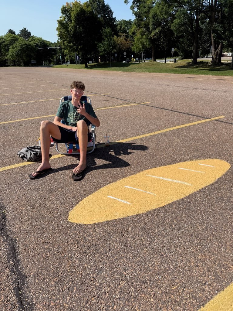 A man in a green shirt and shorts sits on the ground in a parking lot with trees.