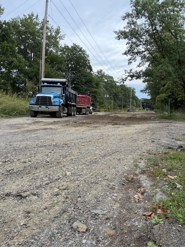 Gravel road with a blue and red dump truck parked in a clearing, surrounded by green trees and shrubs.