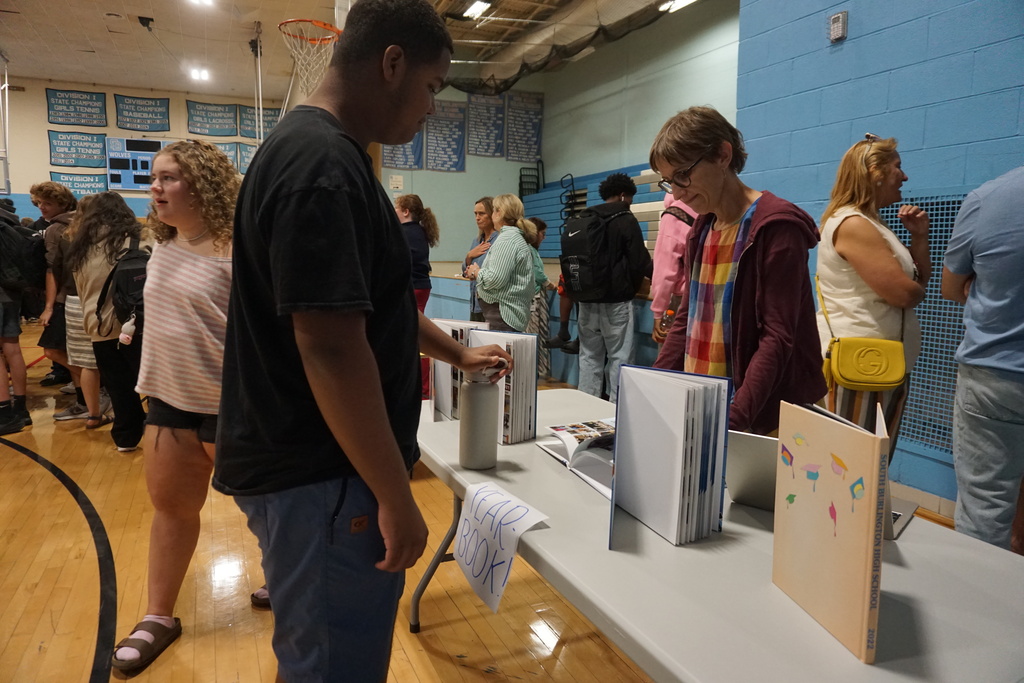 Several people are indoors, standing around a table with books and items on it. The room has wooden floors and blue walls.