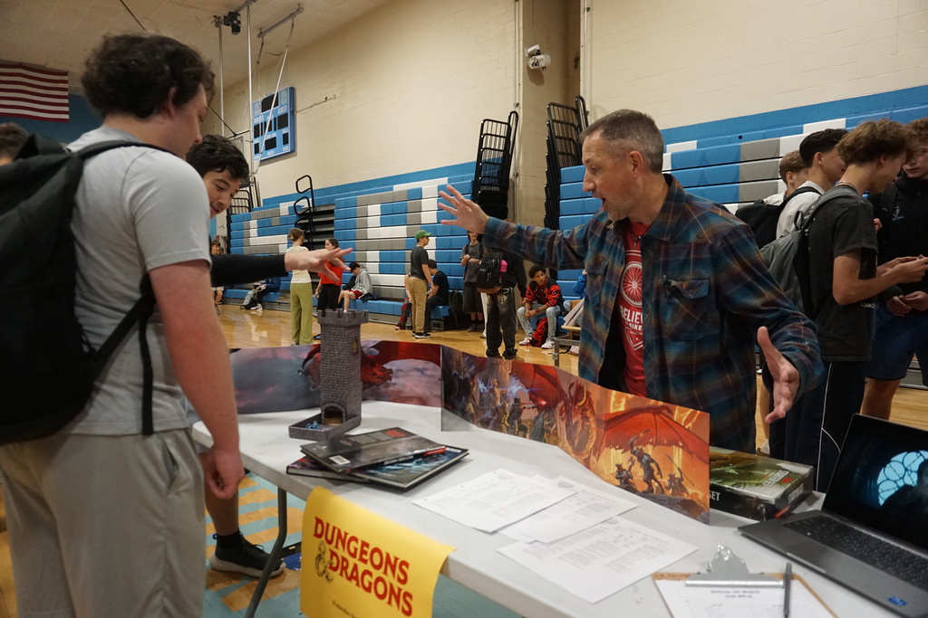 People at a table with Dungeons & Dragons books and a laptop. A man speaks to a youth with a backpack.