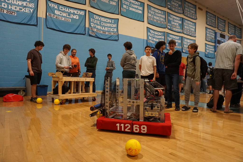 A group of students gather around a robot with yellow balls nearby on a wooden floor.