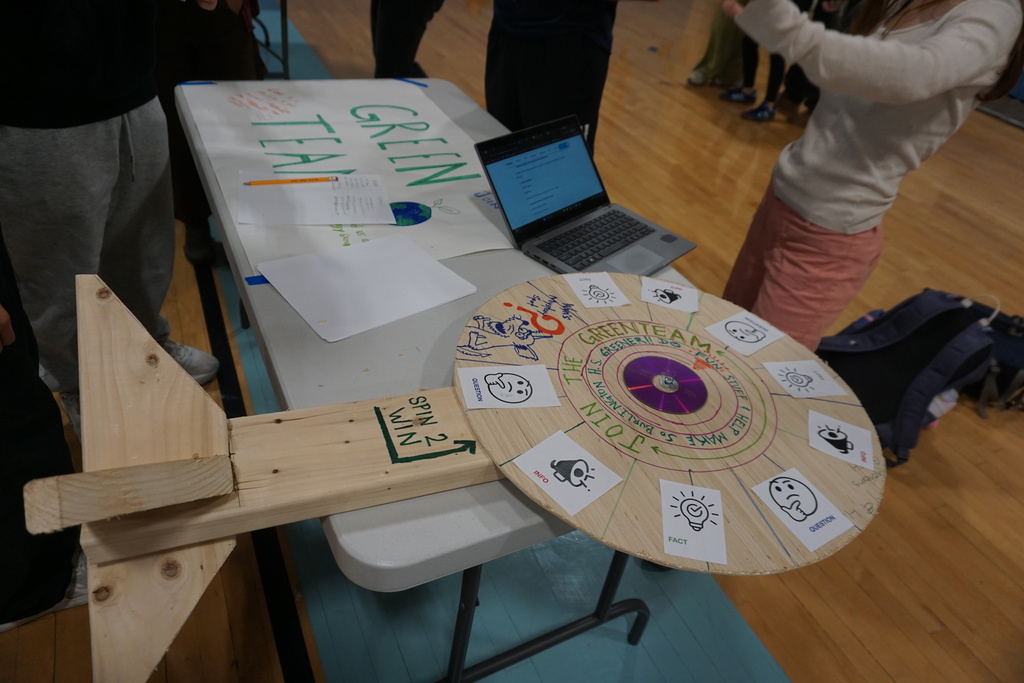 A wooden spinning wheel sits on a table with a laptop and papers. A person stands behind it.
