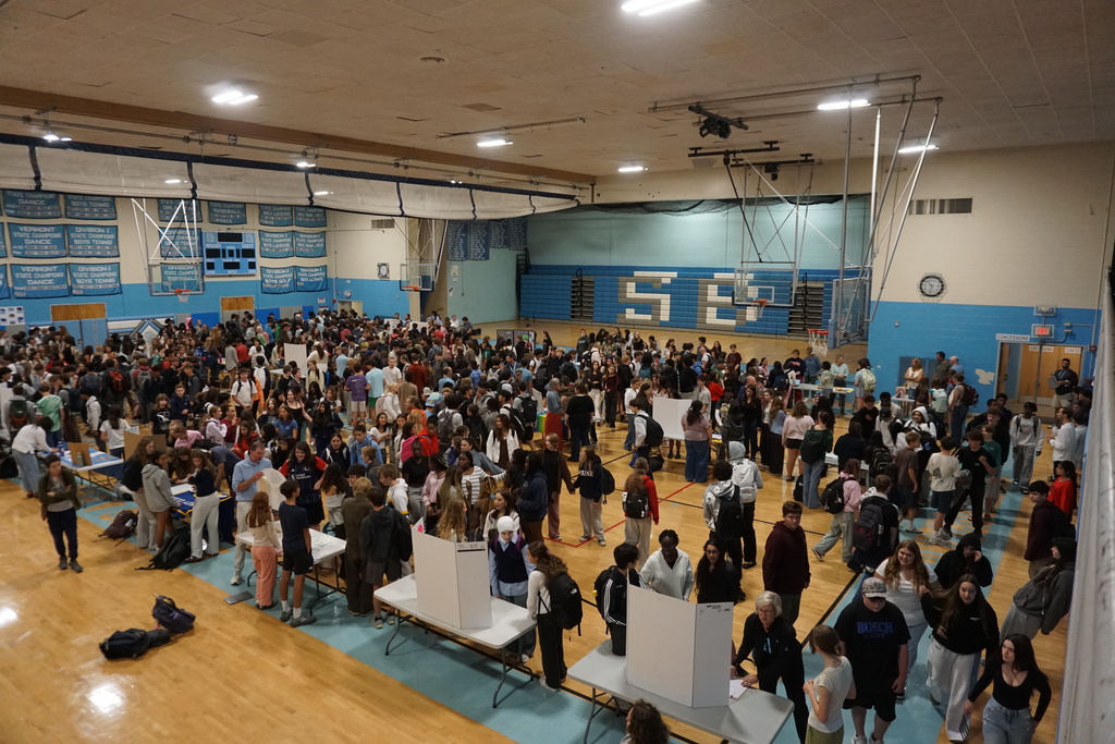 A gymnasium hosts an event with a large group of people standing and walking around, tables, and basketball hoops.