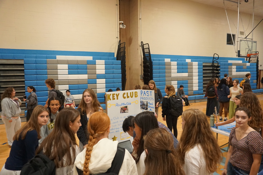 A group of students is gathered in a gym. Some are standing, others are seated, with a large display board in the center.