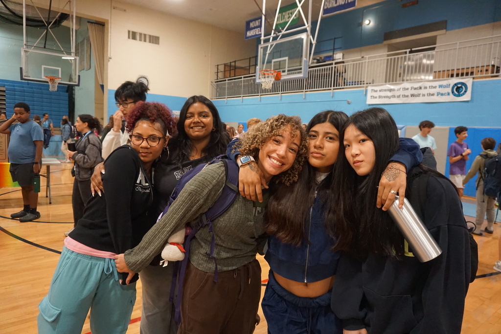 Five women in a gymnasium. Two with curly hair and one with glasses. A basketball hoop is visible in the background.