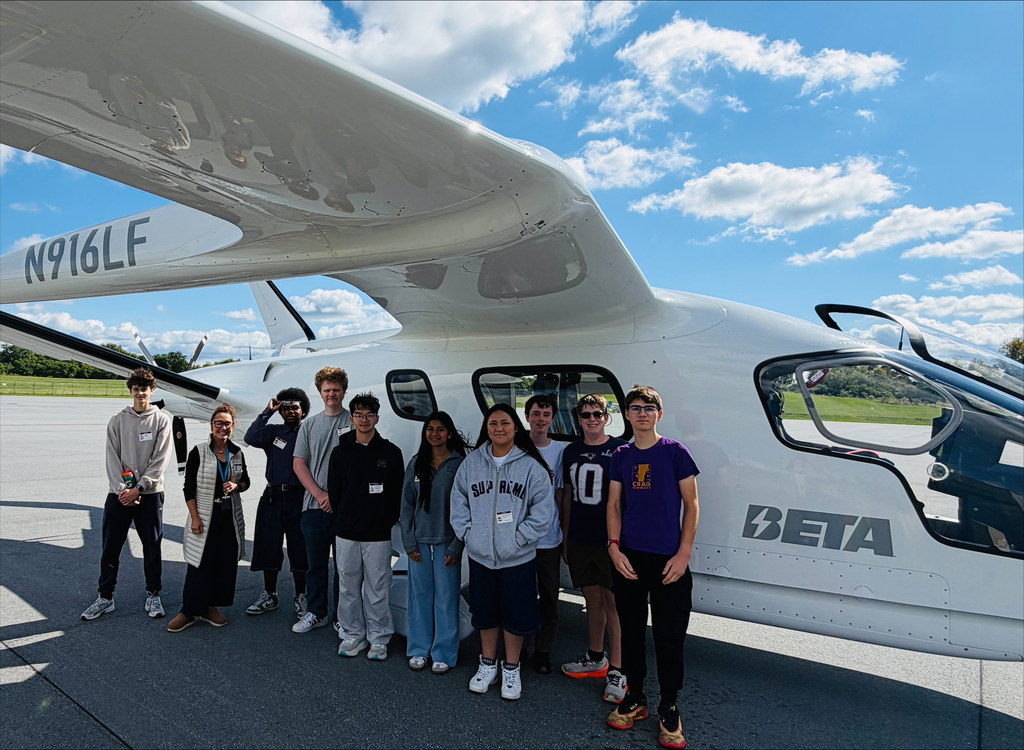 Several individuals stand near a white Beta Aircraft, the model prominently displayed on its side. The sky is blue with some clouds.