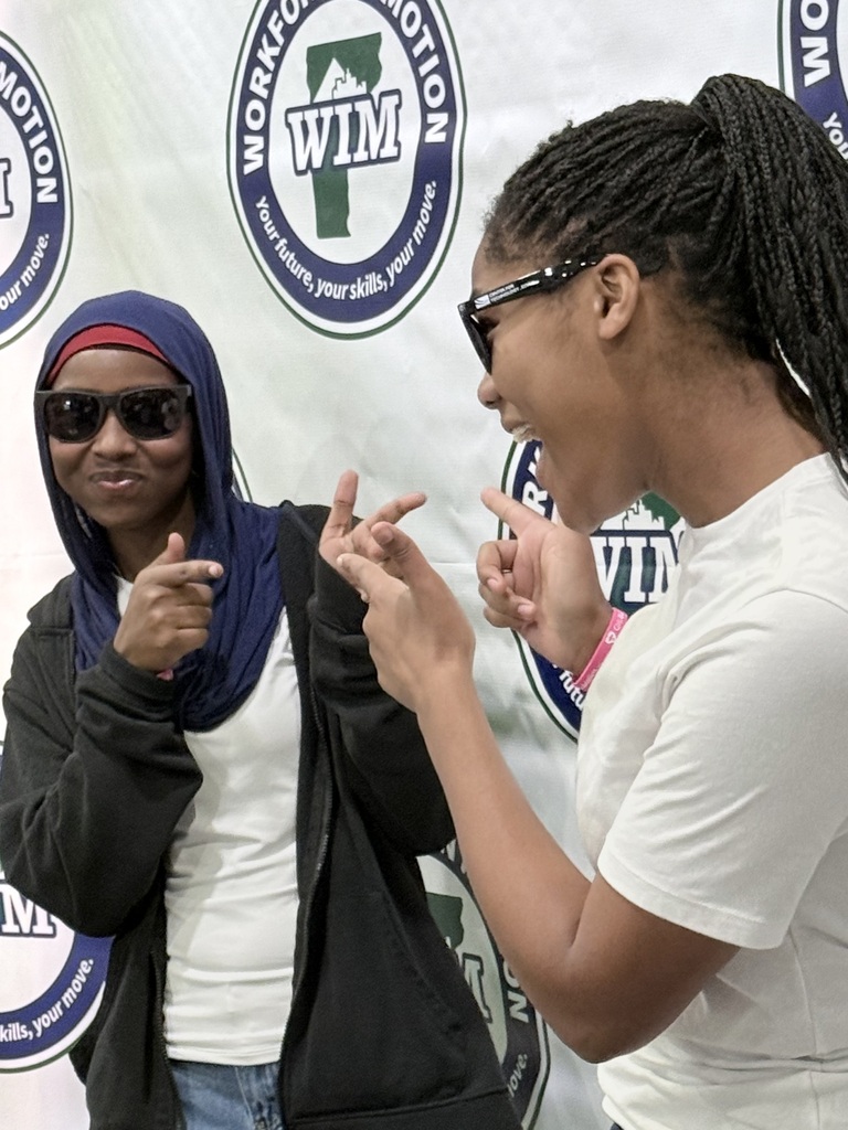 Two women converse and gesture. One wears a blue headscarf, sunglasses, and a black jacket. The other wears a white shirt, glasses, and a pink wristband.