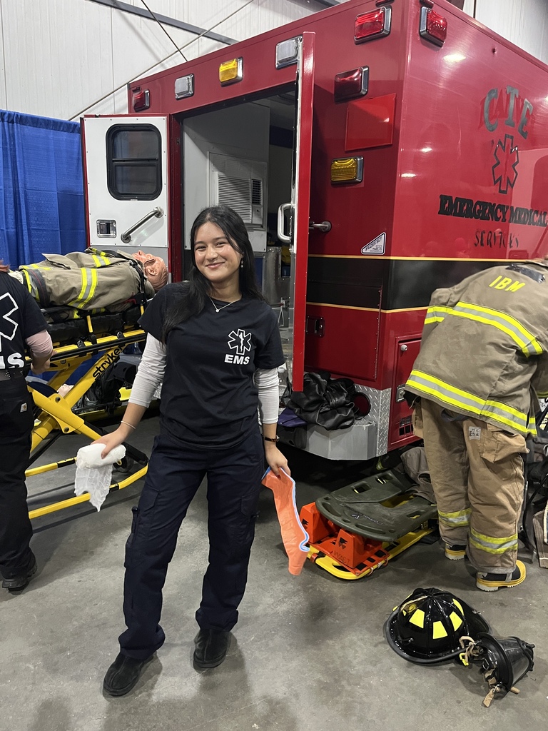 A woman in uniform standing next to an ambulance with an open door, two others in the background.