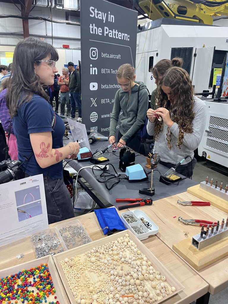 People work on a table with various tools and small objects. One wears a blue shirt and glasses.