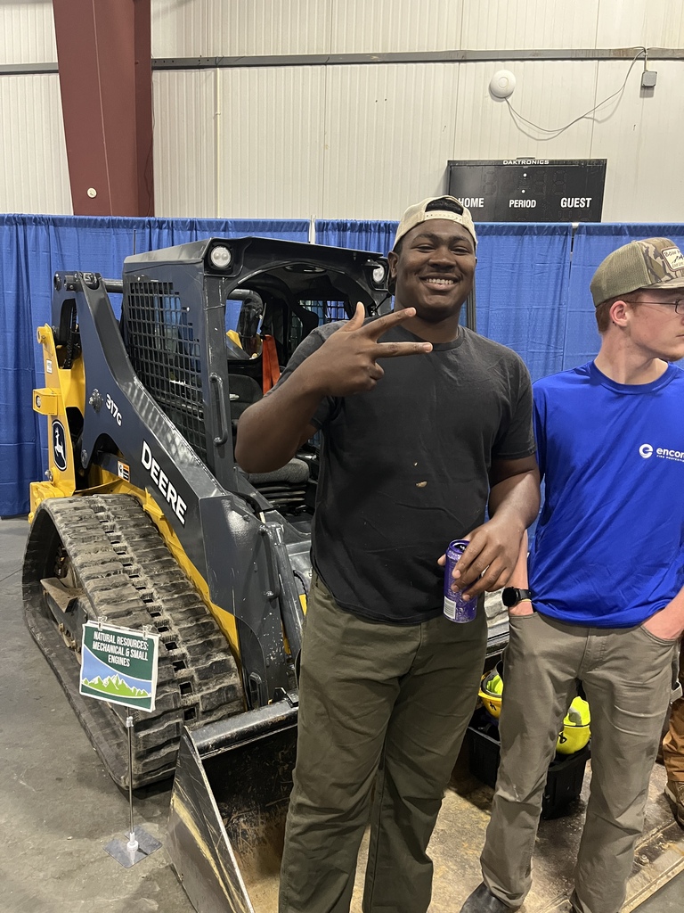 Two men in front of a yellow and black bulldozer, one holding a beverage can, against a blue backdrop.