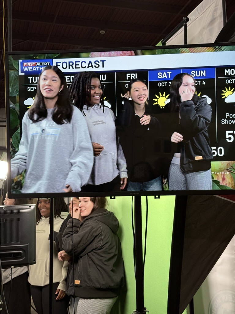 Five individuals standing in front of a screen displaying weather forecast. One woman holds a microphone.