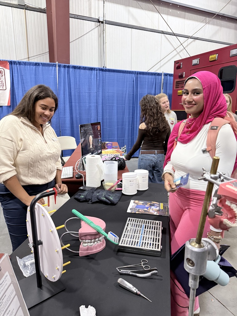 Two women examine a table of dental tools, with a blue backdrop. One woman wears a white sweater, and the other wears a red head scarf.