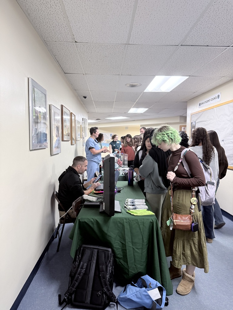 Room with tables, chairs, and people. A man sits at a desk with a computer. Several people stand nearby.