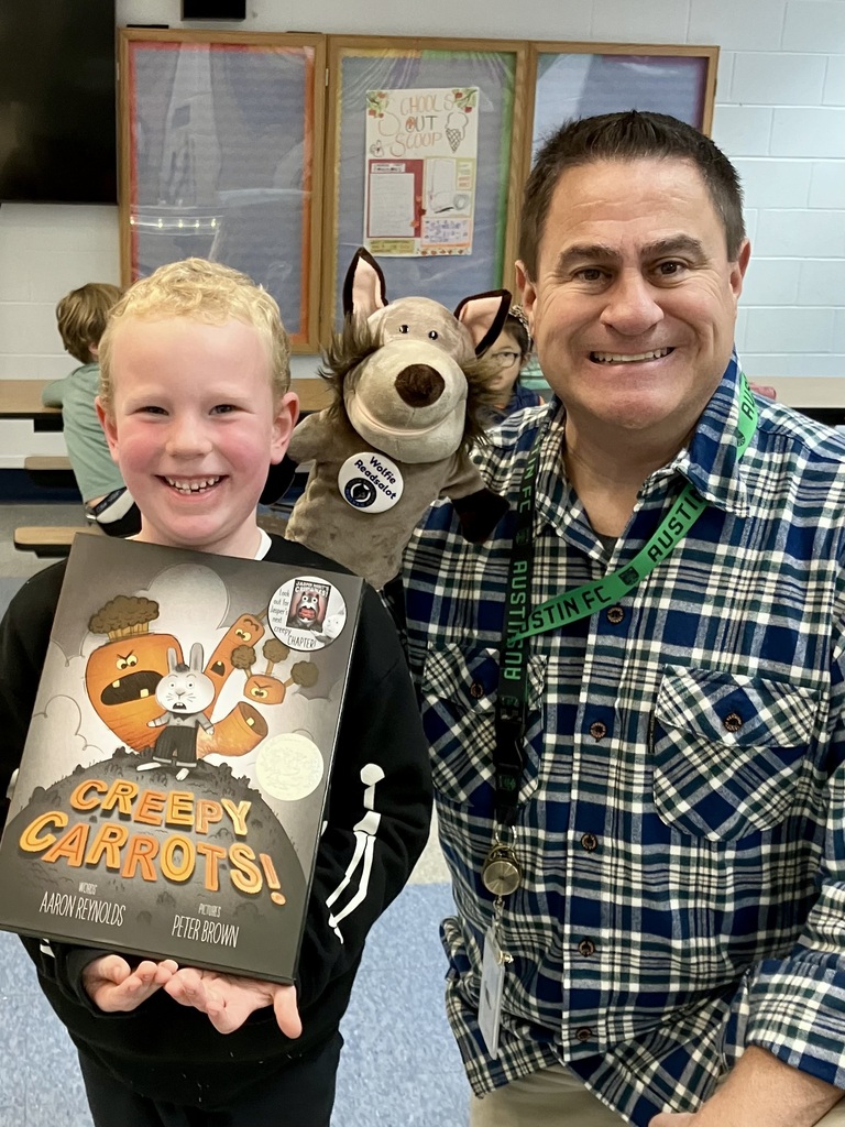 A boy holding a book about creepy carrots stands with a man wearing a green lanyard. Behind them are a stuffed toy and a person.