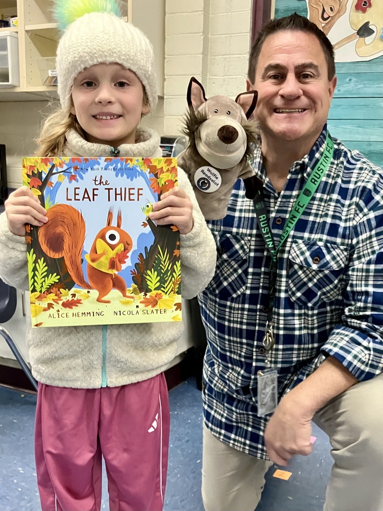 A young girl wearing pink pants holds a book titled "The Leaf Thief" with a squirrel on the cover, next to a man wearing a plaid shirt.