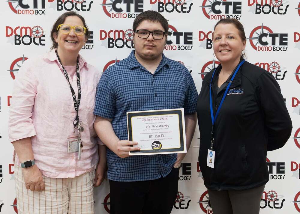 Three people standing next to each other. The person is holding an award. Behind the people is a sign with the words DCMO BOCES, CTE, and image of a circle with a star in the middle.