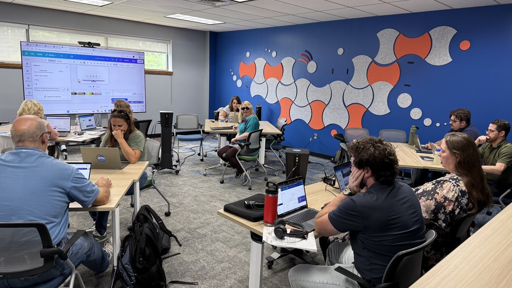 A group of adults participate in a training session in a classroom with laptops at tables. A large display at the front shows a video-editing interface, while a colorful blue, orange, and gray abstract mural decorates the back wall.