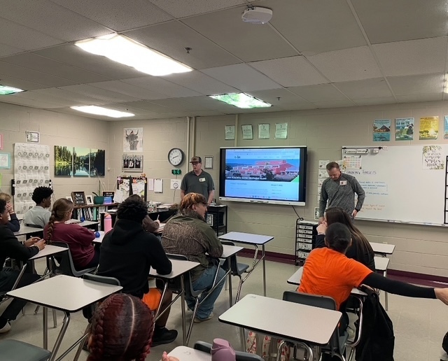 2 adults standing at the front a classroom talking to students sitting in desks