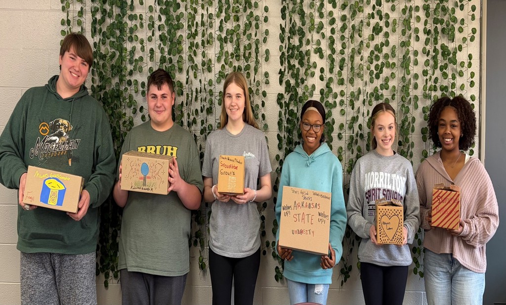 students standing in front of a wall with hanging ivy, eac holding a cardboard box that has been decorated for a packaging  project