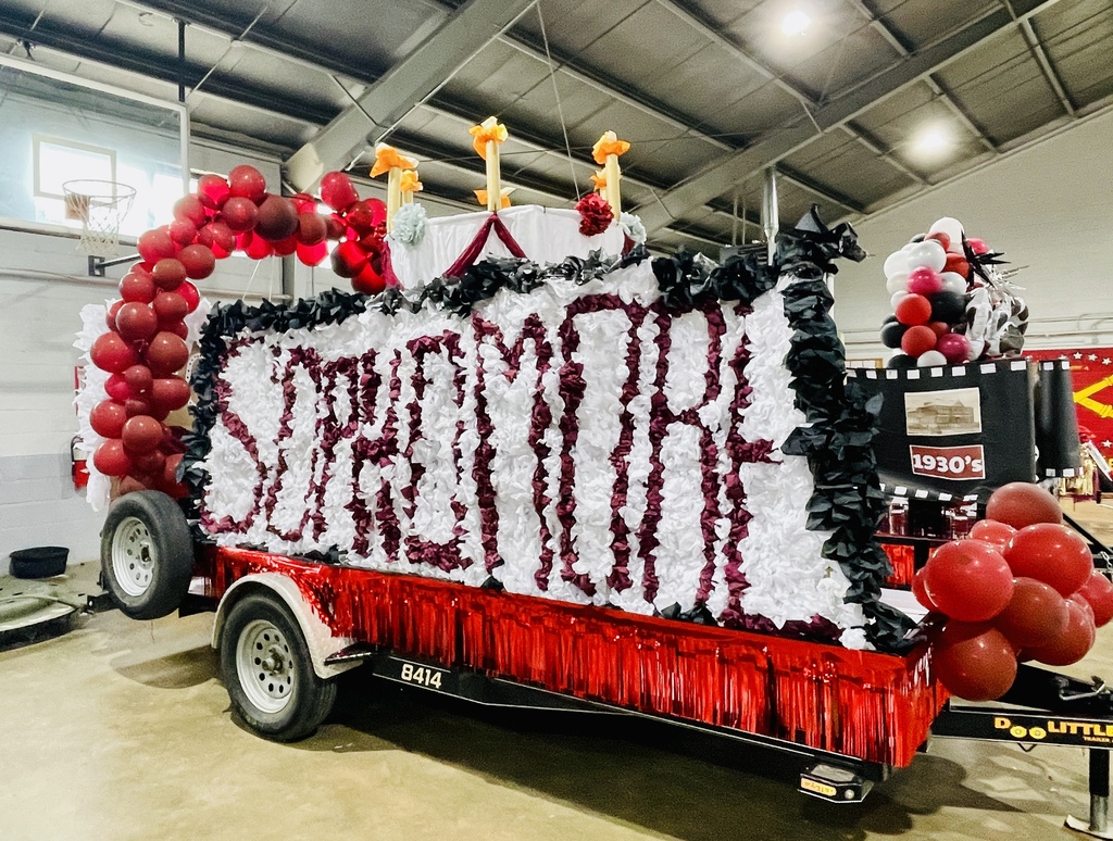 A float with red, white, and black decorations, "SREMORE" written in red, and balloons. It is parked in a large warehouse.